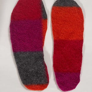 May include: A pair of felted wool shoe insoles featuring a colour-blocked design. The left insole displays horizontal bands of red, magenta, and charcoal grey. The right insole features horizontal bands of orange, red, and magenta.