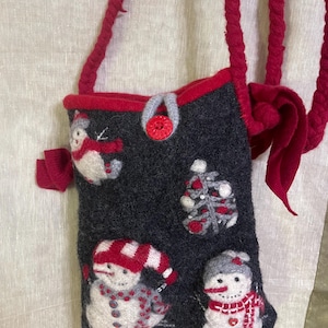 May include: A dark gray felted crossbody bag featuring three white snowmen and a stylized Christmas tree. The bag has a red braided strap, a red button closure, and red trim. The snowmen are decorated with red scarves, hats, and small buttons.