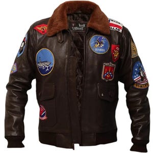 Op de afbeelding: Een bruine leren bomberjack met een bontkraag en meerdere patches. De patches omvatten een marineschip, een "Seabees"-patch, een "Top Gun"-patch en een "USAF"-patch.