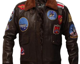 May include: A brown leather bomber jacket with a shearling collar and multiple patches. The patches include a Navy ship, a "Seabees" patch, a "Top Gun" patch, and a "USAF" patch.