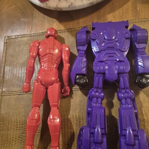 May include: A red plastic action figure of a humanoid character standing next to a larger purple plastic robot action figure. Both figures are shown from the back, displaying molded mechanical details, articulated limbs, and smooth surfaces.