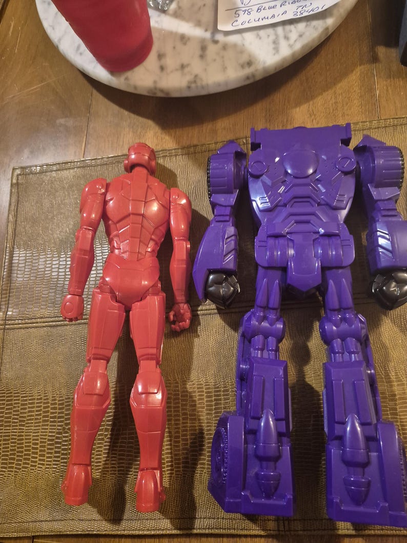 May include: A red plastic action figure of a humanoid character standing next to a larger purple plastic robot action figure. Both figures are shown from the back, displaying molded mechanical details, articulated limbs, and smooth surfaces.