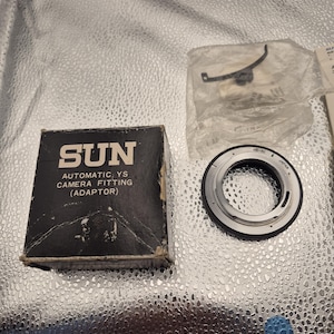 May include: A black and white box with the word "SUN" and the text "AUTOMATIC YS CAMERA FITTING (ADAPTOR)". A silver and black camera lens adapter and a small plastic bag with a metal piece are also visible.