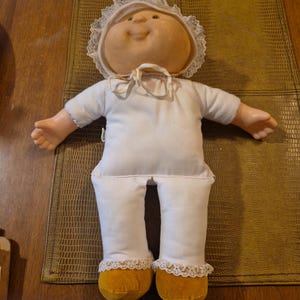 May include: A vintage Cabbage Patch Kid doll, featuring a soft, round face with a smiling expression. The doll is dressed in a white outfit with a lace-trimmed bonnet and yellow shoes. The doll is lying on a brown surface.