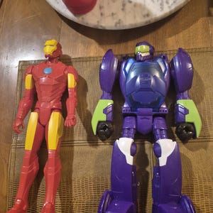 May include: Two plastic action figures. One is a red and yellow Iron Man character with a blue chest arc reactor. The other is a purple, blue, and lime green robot figure with a green visor and a dark blue chest plate featuring a circular emblem.