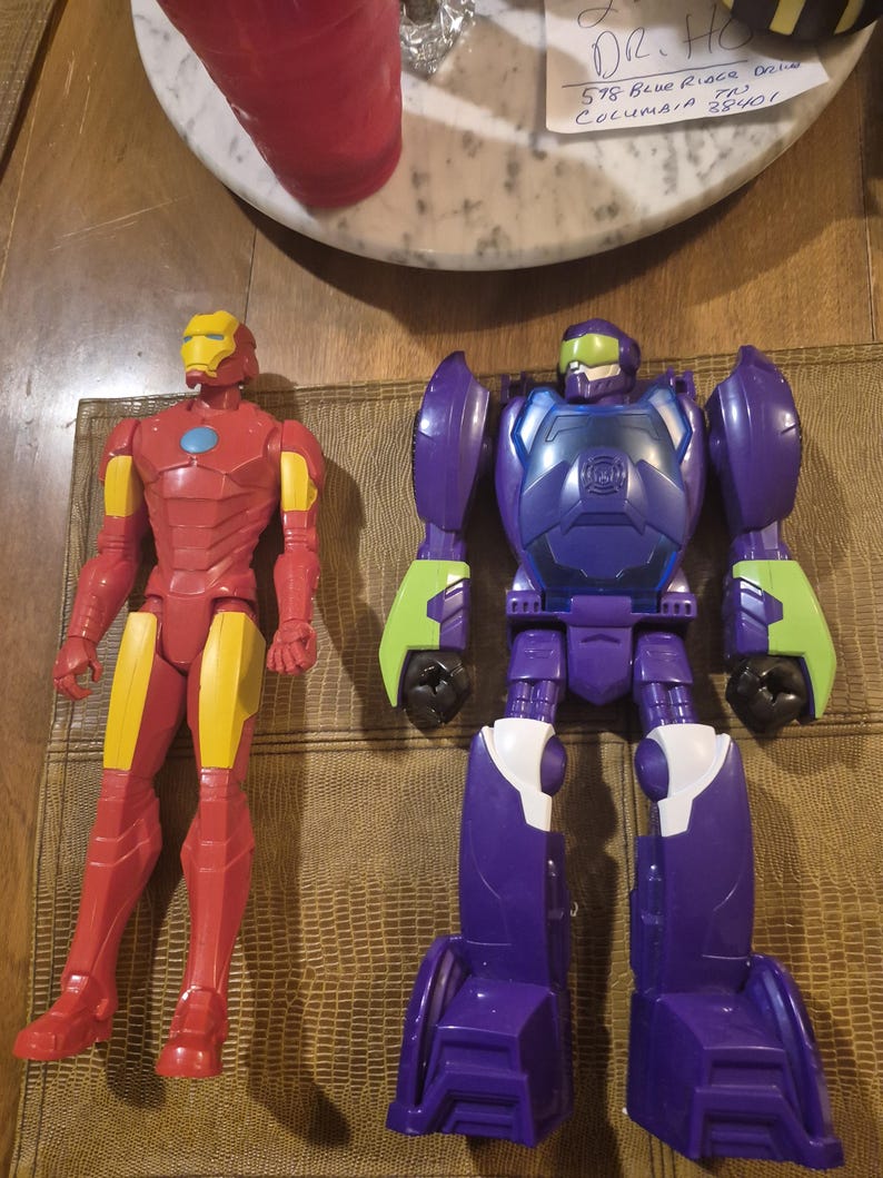 May include: Two plastic action figures. One is a red and yellow Iron Man character with a blue chest arc reactor. The other is a purple, blue, and lime green robot figure with a green visor and a dark blue chest plate featuring a circular emblem.