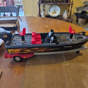 Puede incluir: Barco de pesca de juguete negro y rojo con un remolque. El barco tiene un dise&ntilde;o de llama roja y amarilla y el texto "670 ION" en el lateral. El barco tiene asientos rojos y un volante.