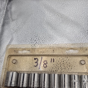 May include: A set of Craftsman sockets in a clear plastic package. The sockets are silver and cylindrical, with the size marked as 3/8". The package is set against a textured, silver background.