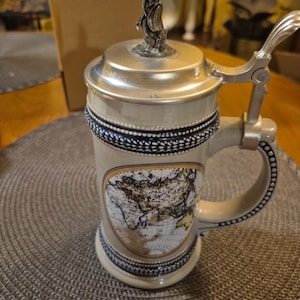May include: A beige ceramic beer stein with a silver lid and handle. The stein features a map of the world and is decorated with blue and black trim. A decorative figure sits atop the lid.