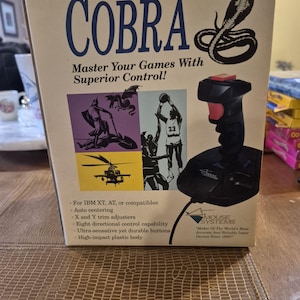 May include: A vintage Cobra joystick game controller box. The box features the word "COBRA" in large blue letters, a cobra illustration, and action figures. The joystick is black with a red top. The box also lists features like auto-centering and durable buttons.