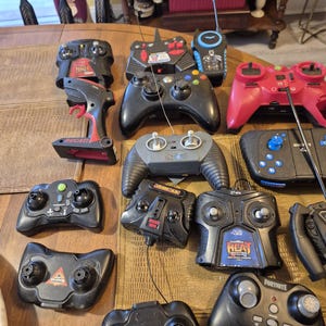 Pu&ograve; includere: Una collezione di vari dispositivi di controllo remoto neri e rossi, inclusi controller di gioco e unit&agrave; portatili. Alcuni controller presentano marchi come "Ducati", "Heat" e "Fortnite". I telecomandi sono disposti su una superficie di legno.