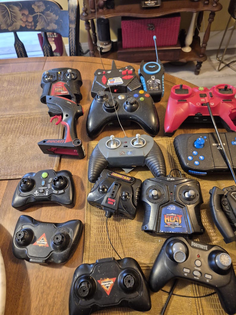 Pu&ograve; includere: Una collezione di vari dispositivi di controllo remoto neri e rossi, inclusi controller di gioco e unit&agrave; portatili. Alcuni controller presentano marchi come "Ducati", "Heat" e "Fortnite". I telecomandi sono disposti su una superficie di legno.