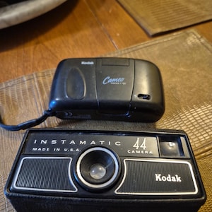May include: Two vintage Kodak cameras. The top camera is black with the word "Cameo" and "Focus Free" printed on it. The bottom camera is a black Instamatic 44 camera with the text "Made in U.S.A." and the Kodak logo.