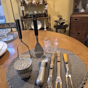 May include: A collection of vintage kitchen utensils, including a potato masher, spatula, ice cream scoop, measuring cup, and forks, arranged on a round placemat. The utensils have black handles and metal components.