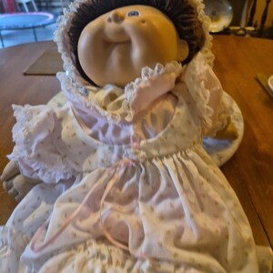May include: A doll with a round, tan face, dimpled cheeks, and brown yarn hair. It wears a white bonnet with lace trim and a white dress featuring a small pink floral pattern, ruffled sleeves, and a pink ribbon tied at the center of the bodice.