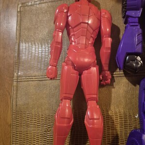 May include: A red plastic action figure shown from the back, featuring a segmented armored design with a helmeted head, articulated limbs, and a smooth finish. Beside it, a portion of a purple and black mechanical toy component is visible.