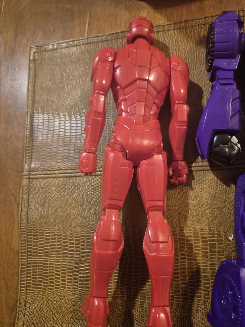 May include: A red plastic action figure shown from the back, featuring a segmented armored design with a helmeted head, articulated limbs, and a smooth finish. Beside it, a portion of a purple and black mechanical toy component is visible.