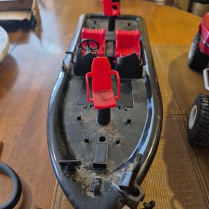 Puede incluir: Barco de juguete de pl&aacute;stico negro con asientos rojos y un volante. El barco tiene un motor y una h&eacute;lice. El barco est&aacute; sobre una superficie de madera.