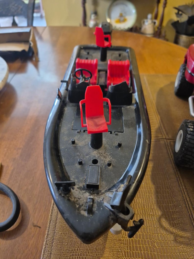 Puede incluir: Barco de juguete de pl&aacute;stico negro con asientos rojos y un volante. El barco tiene un motor y una h&eacute;lice. El barco est&aacute; sobre una superficie de madera.