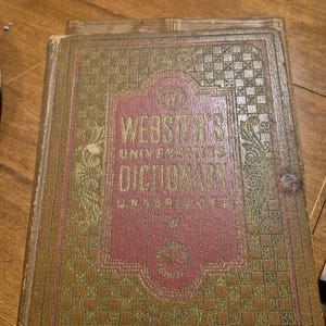 May include: Antique Webster's Unabridged Dictionary with a worn, textured cover. The cover is a combination of brown and red, with gold lettering and decorative patterns. The title is prominently displayed in gold lettering.