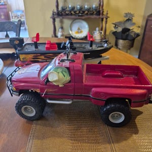 Puede incluir: Un cami&oacute;n de juguete rojo con una caja negra y un pez grande de pl&aacute;stico verde en el asiento del conductor. El cami&oacute;n tiene ruedas negras y un parachoques negro. El cami&oacute;n est&aacute; sentado sobre una superficie marr&oacute;n texturizada.