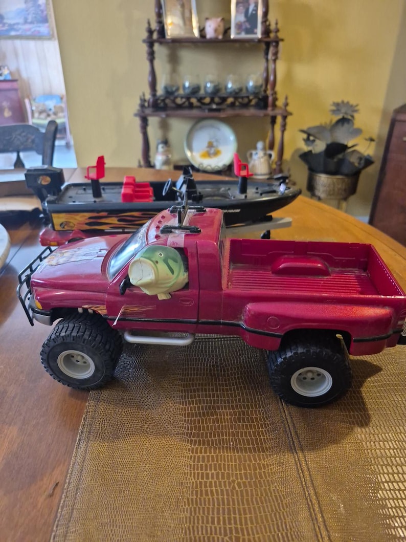 Puede incluir: Un cami&oacute;n de juguete rojo con una caja negra y un pez grande de pl&aacute;stico verde en el asiento del conductor. El cami&oacute;n tiene ruedas negras y un parachoques negro. El cami&oacute;n est&aacute; sentado sobre una superficie marr&oacute;n texturizada.