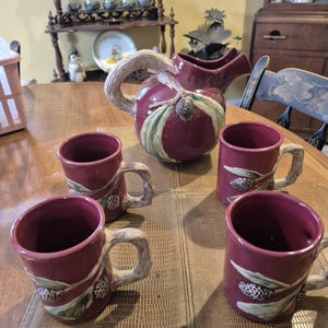 May include: A set of four burgundy mugs and a matching jug with a pine cone and leaf design. The mugs and jug have a rustic, hand-painted look.