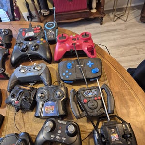 Pu&ograve; includere: Una collezione di vari dispositivi di controllo remoto neri e rossi, tra cui controller di gioco e trasmettitori radio, &egrave; disposta su una superficie di legno. Alcuni controller hanno antenne, pulsanti e joystick. I controller sono di forme e dimensioni diverse.