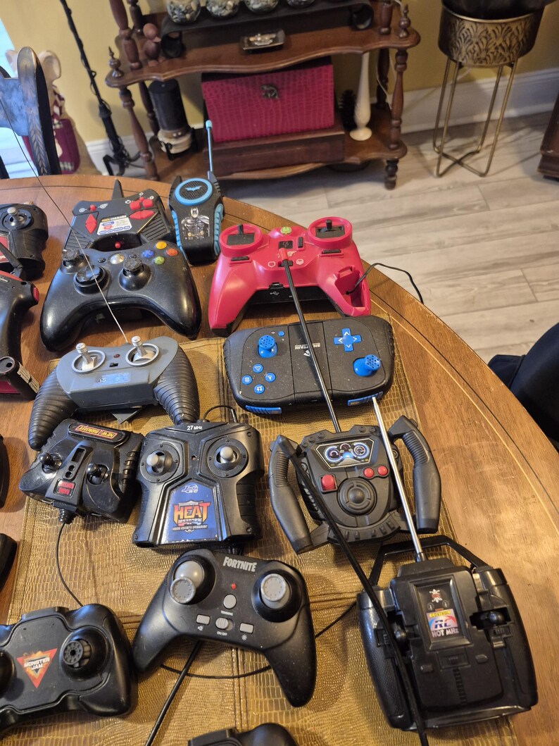 Pu&ograve; includere: Una collezione di vari dispositivi di controllo remoto neri e rossi, tra cui controller di gioco e trasmettitori radio, &egrave; disposta su una superficie di legno. Alcuni controller hanno antenne, pulsanti e joystick. I controller sono di forme e dimensioni diverse.