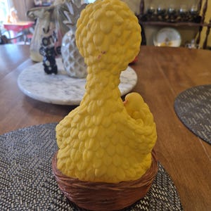 May include: A yellow rubber Big Bird character from Sesame Street sitting in a brown nest. Big Bird is holding a small yellow chick in its right wing.
