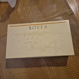 May include: A rectangular box with the word "ROLFS" printed in black. The box is off-white with a distressed appearance and a thin gold stripe. The box is on a brown, textured surface.