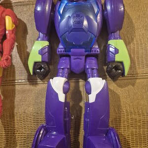 May include: A purple and blue plastic robot action figure with lime green forearm accents and a translucent green visor. The chest plate features a circular emblem, and the legs have white knee details. The figure stands in a vertical position.
