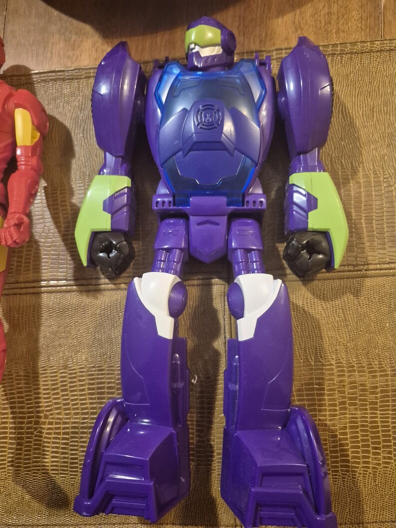 May include: A purple and blue plastic robot action figure with lime green forearm accents and a translucent green visor. The chest plate features a circular emblem, and the legs have white knee details. The figure stands in a vertical position.