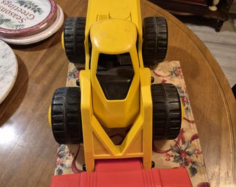 Könnte beinhalten: Ein gelbes Spielzeug-Baufahrzeug mit schwarzen Rädern und einer roten Frontschaufel. Das Spielzeug steht auf einem Läufer mit Blumenmuster auf einem Holztisch. Das Spielzeug ist im Vintage-Stil.
