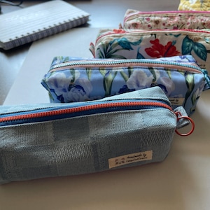 May include: A collection of four rectangular fabric pencil cases arranged in a row. The front case is light blue with a checkered pattern, an orange zip, and a small beige label. Behind it are cases with blue floral, tropical, and floral prints.