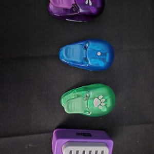 May include: Four different paper punch tools. The top punch is purple and has a dragonfly design. The second punch is blue and has a circle design. The third punch is green and has a paw print design. The bottom punch is purple and has a rectangular design with the word "Micell" printed on it.