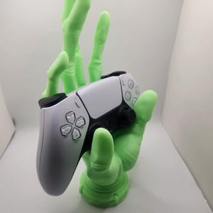 May include: A light green, hand-shaped gaming controller stand holding a white and black controller. The stand has a textured surface and a circular base. The controller has a white face with black accents and buttons.