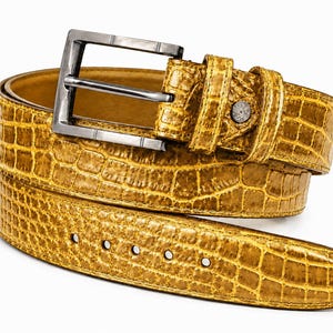 May include: A mustard yellow belt with a textured crocodile skin pattern. The belt features a rectangular gunmetal grey metal buckle and a matching metal loop near the buckle. The strap has five circular holes for adjustment and a pointed end.