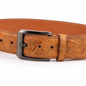 May include: A tan belt featuring a textured crocodile-embossed pattern on the outer surface and a smooth light brown interior lining. The belt is secured with a dark metallic rectangular buckle and includes a matching leather keeper loop.