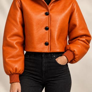 May include: A cropped orange hooded jacket with long sleeves, ribbed cuffs, and three large black buttons down the front. The jacket is worn over a black mock neck top and black denim jeans, creating a layered look with a smooth, solid finish.