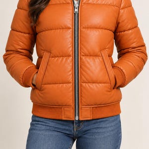 May include: A burnt orange puffer bomber jacket featuring a smooth, leather-like finish, horizontal quilted stitching, a ribbed collar, ribbed cuffs, and a central silver-toned zipper closure with a black trim. The jacket includes two side pockets.