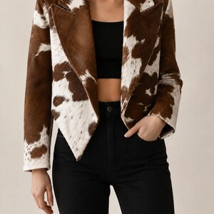 May include: A cropped blazer featuring a brown and white cow print pattern with a notched lapel collar and long sleeves. The jacket is worn over a black crop top and black high-waisted trousers, creating a structured and layered appearance.