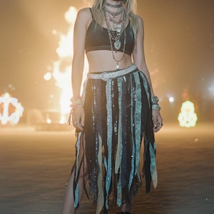 May include: A woman wearing steampunk-inspired attire. She is wearing a black crop top, layered necklaces, a fringed skirt with shimmering accents, and platform boots. Goggles rest on her forehead. The background is a warm, hazy scene.
