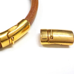 May include: A gold-colored magnetic clasp, detached from a brown leather cord. The clasp is a rounded rectangular shape with a curved design. The leather cord is a light brown color. The background is white.