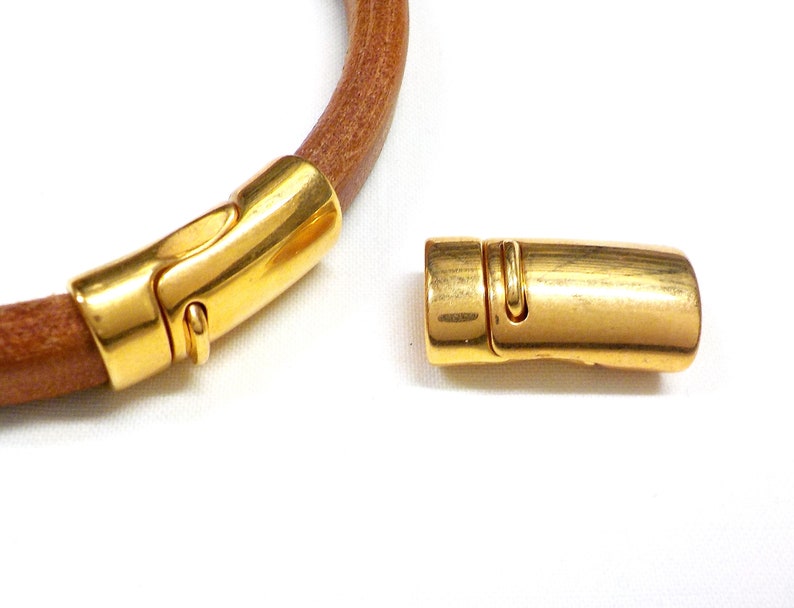 May include: A gold-colored magnetic clasp, detached from a brown leather cord. The clasp is a rounded rectangular shape with a curved design. The leather cord is a light brown color. The background is white.