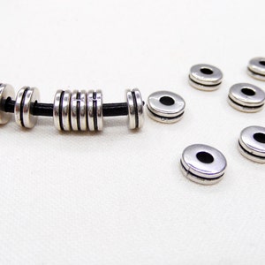 Op de afbeelding: Zilveren metalen kralen met een zwart leren koord. De kralen zijn rond en hebben een gladde afwerking. Er zijn meerdere kralen op het koord en meerdere losse kralen.