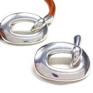 May include: Two silver oval-shaped clasps with a smooth, shiny finish. The clasps have a unique design with a small, rounded protrusion on one side.