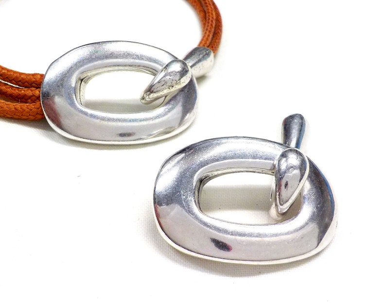 May include: Two silver oval-shaped clasps with a smooth, shiny finish. The clasps have a unique design with a small, rounded protrusion on one side.