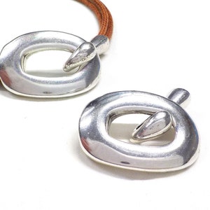 May include: Two silver metal oval-shaped clasps with a rounded, smooth, and slightly raised center. The clasps are designed to be used as a closure for a leather cord or strap.