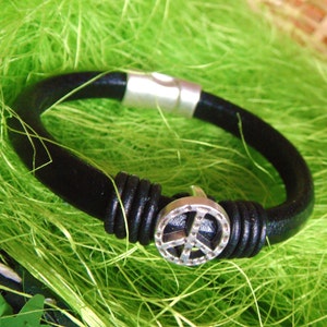 May include: Black leather bracelet with a silver peace sign charm and a magnetic clasp. The bracelet is set against a bright green background, highlighting the contrast between the dark leather and the shiny metal.